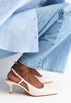 Donkerhuidige persoon die lichtblauwe spijkerbroek en off-white lage slingback schoenen met een gestructureerd patroon draagt, geposeerd tegen een witte achtergrond.