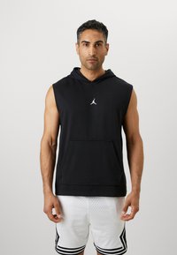 Svart ermeløs hettegenser med frontlomme, med en liten hvit logo på brystet. Kombinert med hvite mesh-shorts.