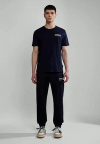 Navy modrá bavlněná tričko s krátkým rukávem a logem na hrudi, spárované se shodnými navy modrými tepláky. Boty jsou bílé s černými akcenty.