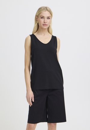 Szőke nő arany fülbevalóval, ujjatlan fekete felsőben és fekete, térdig érő rövidnadrágban, egyszínű világos háttér előtt állva.