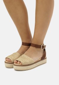 Espadrilles à talon plateforme en cuir marron avec une empeigne tissée beige, semelle texturée et bride chevillère. Design ouvert avec des détails de boucle.