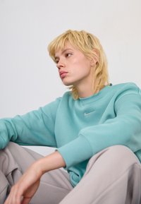 Una persona joven con el cabello corto y rubio lleva un sudadera de Nike color teal y pantalones beige, sentada con los brazos descansando sobre las rodillas contra un fondo sencillo.