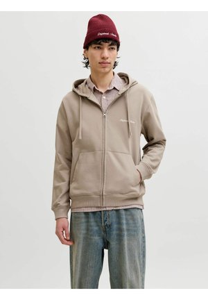 Młody mężczyzna ubrany w beżową bluzę na zamek błyskawiczny na koszulę w kratę, luźne niebieskie dżinsy i czerwony beanie na jednolitym szarym tle.