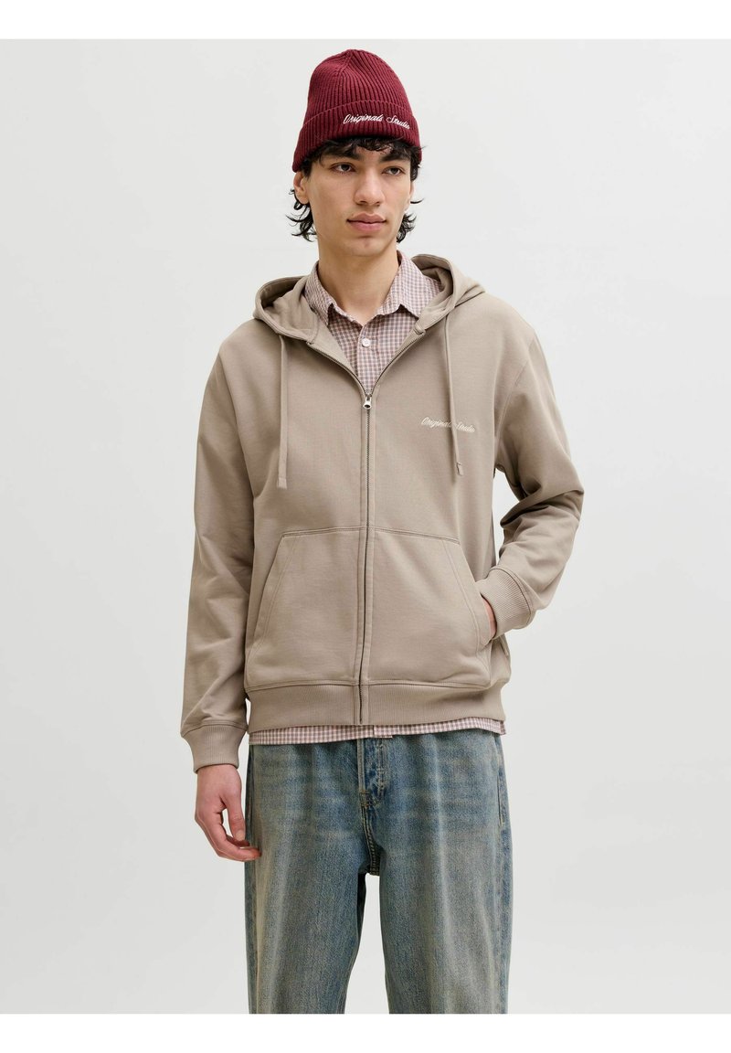 Jeune homme portant un sweat à capuche beige zippé sur une chemise à carreaux, un jean bleu ample et un bonnet rouge sur un fond gris uni.