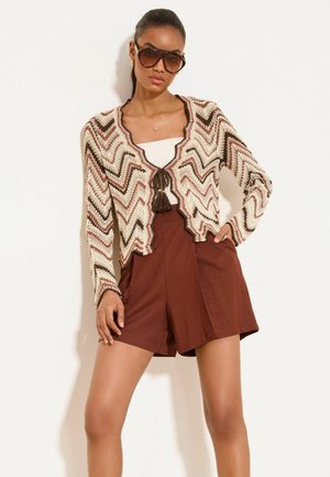 Kobieta nosząca duże okulary przeciwsłoneczne, sweter z dzianiny z wzorem w jodełkę i wiązaniami z chwostami, beżowy top i brązowe, wysokie szorty, pozująca z ręką na biodrze.