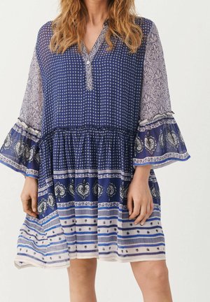 Niebiesko-biała sukienka o długości do kolan z dzwonkowatymi rękawami i luźnym krojem, noszona przez osobę o falowanych blond włosach.