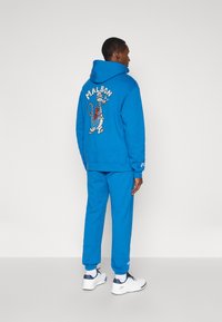 Zila kapucēta džemperis ar grafisku modeli aizmugurē, apvienots ar atbilstošām šaurām biksēm. Tērps ir aprīkots ar ribotām aprocēm un logotipa akcentiem.