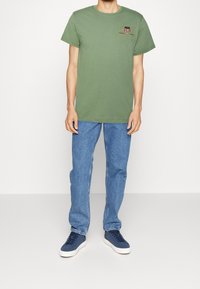 Camiseta de algodón verde oliva con un gráfico en el pecho, combinada con jeans de mezclilla azul claro y zapatillas navy con suelas blancas.