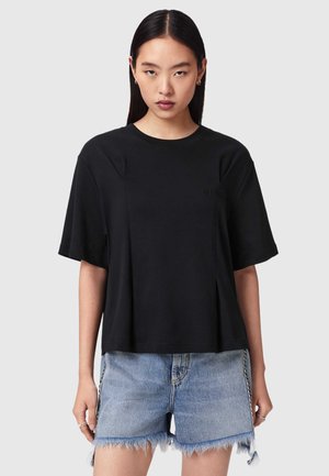 Kvinde med lige mørkt hår iført en enkel sort T-shirt med korte ærmer og slidte denimshorts med frynser, stående foran en lysegrå baggrund.