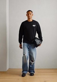 Schwarzer Sweatshirt mit weißem Logo, hellblaue, gewaschene Jeans mit fransigen Rändern und schwarze Ledertasche, kombiniert mit schwarzen Schuhen auf einem Holzboden.