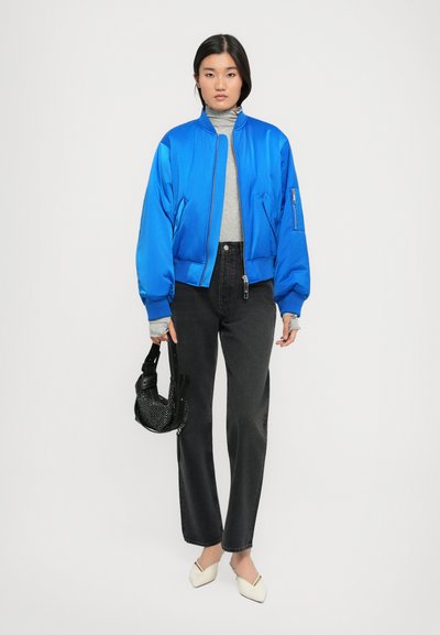 Blaue Satin-Bomberjacke mit gerippten Bündchen und Kragen, grauer Rollkragenpullover, schwarze hochtaillierte Jeans und weiße Slipper, hält eine schwarze Tasche.