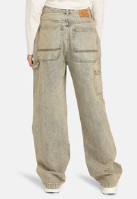 Persona que lleva unos pantalones cargo holgados de lavado claro con grandes bolsillos traseros y laterales, combinados con zapatillas blancas y una camiseta blanca.
