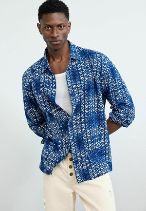 Mand iført blå mønstret skjorte med knapper over hvid tanktop og beige bukser, stående med hænderne bag ryggen mod en hvid baggrund.