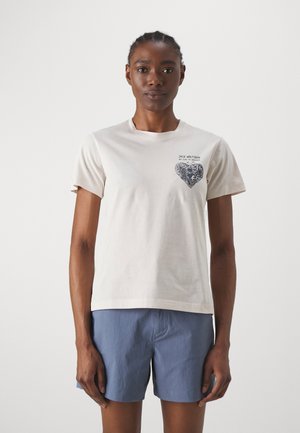 T-shirt in cotone beige con maniche corte, caratterizzato da una grafica a forma di cuore blu scuro sul lato sinistro del petto, scollo rotondo e vestibilità morbida.