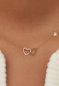Goldene Halskette mit zwei ineinandergreifenden Herzanhängern, die ein schlichtes Design und eine dünne Kette aufweist. Ein kleiner Herzanhänger ist ebenfalls angebracht.