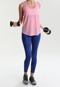 Rosa ärmlös tanktopp, blå mönstrade leggings och svarta träningsskor. Håller två svarta hantlar, vilket visar en sportig fitnesslook.