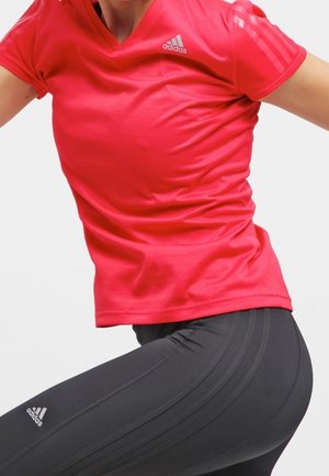 Person trägt ein knallrotes Adidas-Shirt und schwarze Adidas-Leggings, posiert in Bewegung vor weißem Hintergrund.