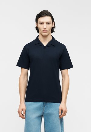 Young man with dark hair wears a short-sleeve black polo shirt and light blue jeans, standing against a plain white background.