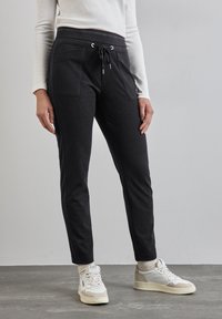 Pantalones deportivos negros con cintura ajustable, dos bolsillos frontales, diseño de tobillo entallado, y combinados con zapatillas beige y blancas.