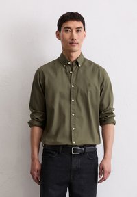 Olijfgroene button-up shirt met opgerolde mouwen, witte knopen en een klein geborduurd logo. Gepaard met donkere jeans en een zwarte riem.