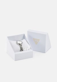 Zilveren polshorloge met een ronde wijzerplaat, metalen schakelarmband, gepresenteerd in een gestructureerde witte doos met een gouden logo. Zachte witte kussen binnenin.