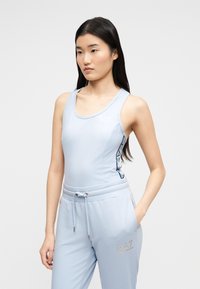 Γυναίκα με μακριά σκούρα μαλλιά, φοράει αμάνικη μπλούζα ανοιχτού γαλάζιου και ταιριαστό παντελόνι jogger, στέκεται με το ένα χέρι στην τσέπη.