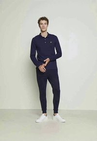 Camisa polo de manga longa azul-escura com colarinho e um pequeno logótipo amarelo, combinada com calças slim-fit da mesma cor e ténis brancos.