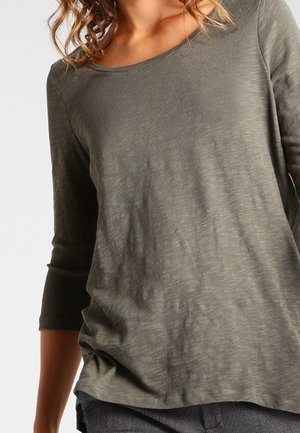 Kobieta ubrana w oliwkowozieloną koszulę z rękawem do łokcia, o zaokrąglonym dekolcie i luźnym kroju.