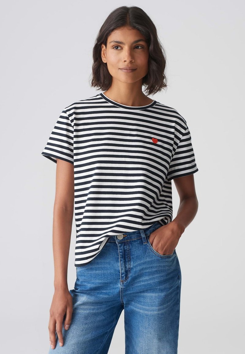 Zwart-wit gestreepte kortemouwen t-shirt met een ronde hals en een klein rood hartdetail op de borst, gecombineerd met blauwe spijkerbroek.