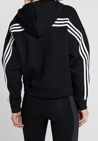 Felpa nera con strisce bianche lungo le spalle, dotata di cappuccio con cordini e vestibilità comoda. Abbinata a leggings neri con motivi a taglio.