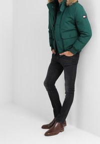 Giacca a vento verde con cappuccio foderato in pelliccia, due tasche frontali, abbinata a jeans scuri e stivaletti marroni. Texture liscia, design casual.