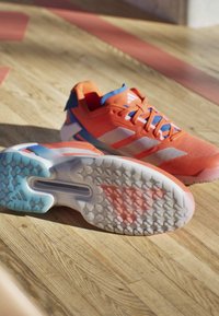 Coppia di scarpe sportive arancioni e bianche con accenti blu su un pavimento in legno, una mostra la suola strutturata e il logo Adidas.