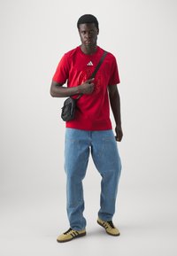 Red Adidas t-shirt with logo, loose light blue jeans, and yellow sneakers with black stripes. Black messenger bag worn across the body.