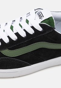 Fekete velúr sneaker fehér fűzős, zöld díszítéssel és zöld gumis oldalsó csíkkal. Párnázott gallérral és fehér talppal rendelkezik.