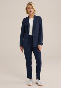 Mörkblå skräddarsydd kostym med enbladsblazer och smala byxor, matchad med en vit topp och avslappnade vita sneakers.