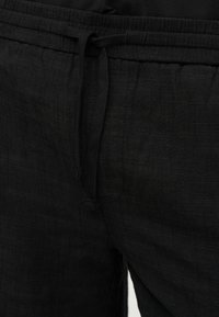 Pantalon noir en tissu texturé avec cordon à la taille, montrant un gros plan de la partie avant de la taille.