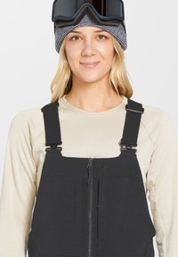 Schwarze Skihosen mit verstellbaren Trägern, Reißverschluss vorne und zwei Fronttaschen. Getragen über einem cremefarbenen Langarmoberteil und einer Strickmütze.