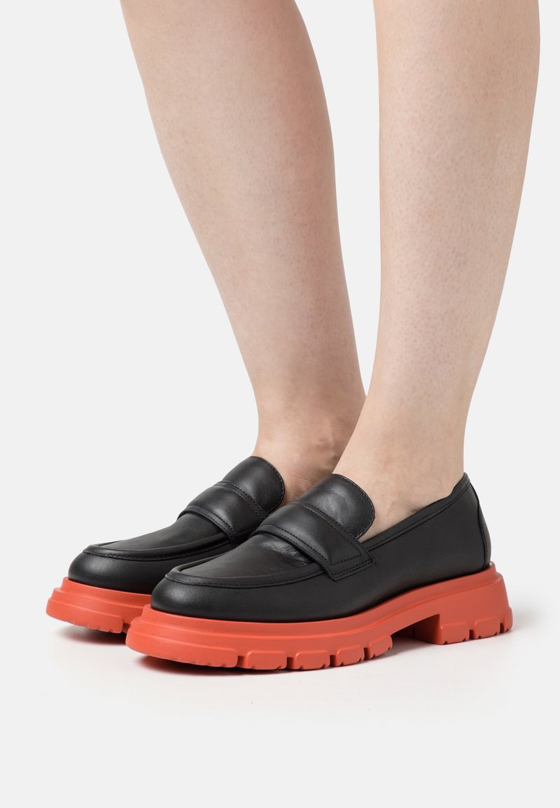 Zwarte leren loafers met een dikke oranje rubberen zool, voorzien van een band over de bovenkant en een gladde, matte afwerking. Gestructureerde accent op de zool.