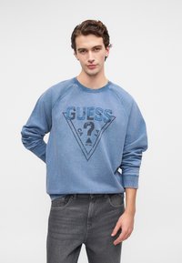 Joven con pelo rizado corto que lleva una sudadera azul de GUESS y jeans grises, de pie contra un fondo blanco liso.