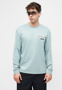 Νεαρός άνδρας με ανοιχτό γαλάζιο πουκάμισο μακρυμάνικο με τσέπη στο στήθος που κλείνει με φερμουάρ και σκούρα γυαλιά ηλίου, στέκεται μπροστά σε ένα απλό λευκό φόντο.