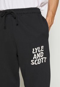 Czarne dresy z elastycznym paskiem i sznurkiem do regulacji. Lewa nogawka ozdobiona białym haftem z napisem "LYLE AND SCOTT."