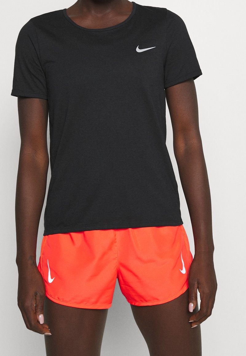 En person klädd i en svart Nike T-shirt och ljus orange Nike träningsshorts, stående framför en enkel ljus bakgrund.