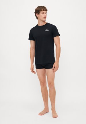 Schwarzes Baumwoll-T-Shirt mit kurzen Ärmeln, mit einem kleinen Logo auf der Brust. Getragen mit passendem schwarzen Slips, Modell steht barfuß.