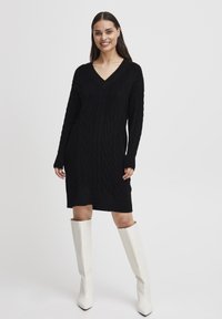 Abito nero in maglia con motivi a treccia, scollatura a V, maniche lunghe e orlo a costine. Indossato con stivali bianchi al ginocchio con punta affusolata.