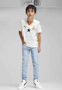 Balts krekls ar Ganas nacionālās komandas dizainu, ar melno zvaigzni, daudzkrāsainu apkakli, pārī ar gaiši ziliem džinsiem un baltiem sporta apaviem.