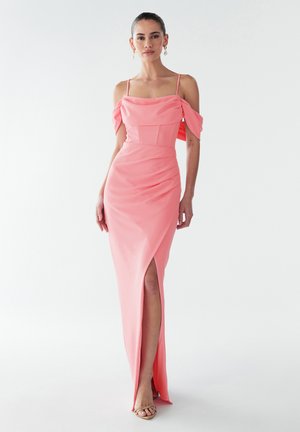 Moteris rožinės spalvos su pečių iškirpte suknelėje su spaghetti dirželiais, aukštai iki šlaunies perplėšimu ir kreminės spalvos atvirais aukštakulniais, prieš balto fono.