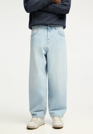 Persona con vaqueros anchos de pierna color azul claro, zapatillas blancas y una camiseta oscura de manga larga, de pie con los brazos cruzados.