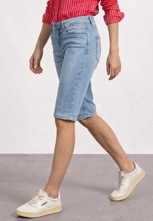 Person, die hellblaue Jeansshorts, ein rotes langärmeliges gestreiftes Hemd und weiße Sneakers auf grauem Boden trägt.