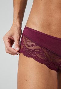 Bordeaux kanten slipjes met een doorschijnende kanten afwerking langs de zijkanten. De stof heeft een gladde textuur die de delicate kanten details benadrukt.