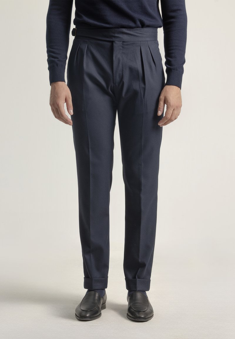 Hombre vestido con pantalones azul marino con pliegues, mocasines negros y una camiseta de manga larga azul marino, de pie contra un fondo claro y uniforme.